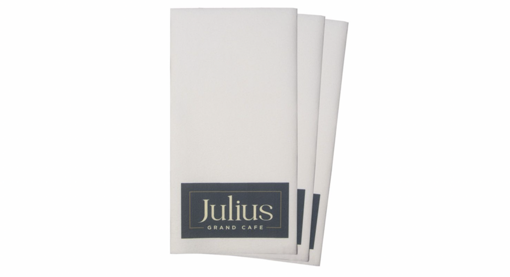 Weiße, bedruckte Airlaid-Servietten mit elegantem dunkelblau-goldenem Logoaufdruck „Julius Grand Café“. Hochwertige Präsentation für Gastronomie, stilvolle Cafés und gehobene Restaurants, funktional und zugleich repräsentativ.