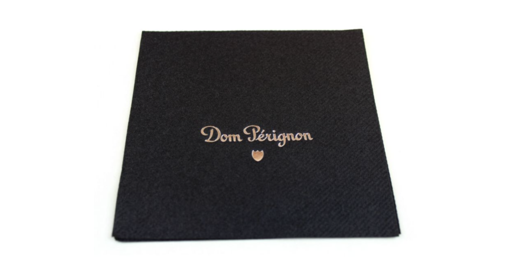 Schwarze, individuelle Airlaid-Serviette mit edlem, goldfarbenem Aufdruck „Dom Pérignon“ und kleinem Logo-Symbol. Hochwertiges Design für gehobene Gastronomie, Bars und exklusive Events, stilvoll und elegant präsentiert.