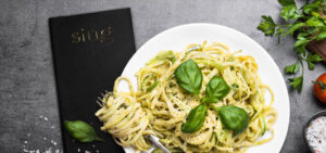 Eine individuelle Airlaid-Serviette liegt auf einem feinen Restauranttisch. Die schwarze Airlaid-Serviette ist mit goldener Schrift bedruckt. Auf dem Tisch steht zudem ein weißer Teller mit Pasta.